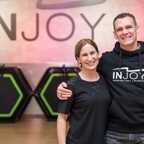 Zwei lächelnde Menschen im INJOY Fitnessstudio vor Trampolinen und Trainingsgeräten.