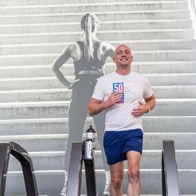 Mann joggt lachend auf Laufband im Fitnessstudio vor großem Motiv mit Treppe und Schriftzug.