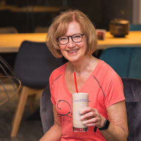 Ältere Frau mit Brille und sportlichem Shirt genießt lächelnd einen gesunden Proteinshake.
